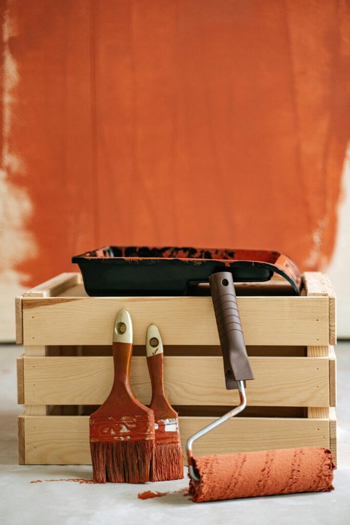Wooden crate with paintbrushes and roller against a partially painted wall.